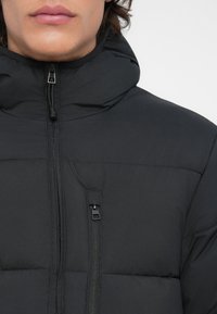 Primo piano di una persona che indossa una giacca invernale nera imbottita con colletto alto e due tasche verticali con zip.