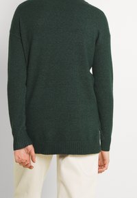 Dunkelgrüner Strickpullover mit lässiger Passform, gerippten Bündchen und Saum. Verfügt über Seitenschlitze und eine weiche Textur. Sichtbar über einer crèmefarbenen Hose getragen.