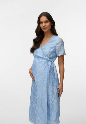 Kobieta z długimi ciemnymi włosami w teksturowanej, jasnoniebieskiej sukience kopertowej delikatnie trzyma ciążowy brzuch, stojąc na tle jednolitego białego tła.