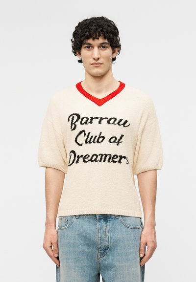 Jeune homme aux cheveux courts, bouclés et foncés, portant un pull en maille crème avec un col en V rouge et le texte noir "Barrow Club of Dreamers" ainsi qu'un jean bleu clair.