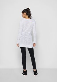 Pull en tricot blanc avec un tissage lâche et ouvert, des manches longues et un dos légèrement allongé, associé à un legging noir et des baskets noires.