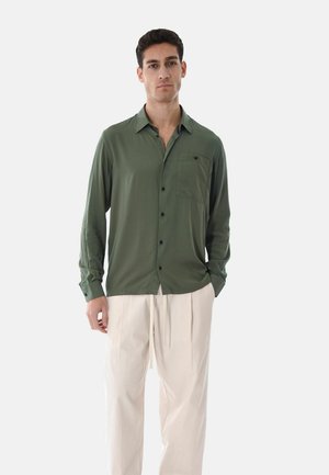 Uomo in piedi che indossa una camicia verde oliva chiara con bottoni e taschino sul petto, abbinata a pantaloni bianchi a gamba larga, su sfondo neutro.