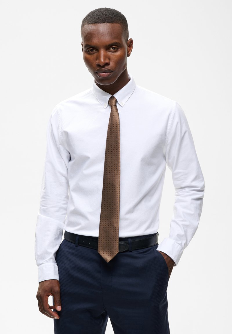 Jeune homme portant une chemise blanche, une cravate marron, un pantalon bleu marine et une ceinture noire, debout avec une main dans la poche devant un fond uni.