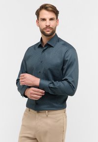 Camicia button-up color verde scuro con un sottile motivo a pois, maniche lunghe e polsini con un bottone, abbinata a pantaloni beige.