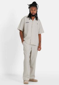 Camisa de manga corta a rayas y pantalones a juego hechos de una tela ligera en tonos beige y crema. Presenta bordados y bolsillos laterales. Sandalias puestas.