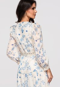 Femme aux longs cheveux foncés portant une robe blanche à motifs floraux bleus, avec des manches bouffantes et un galon en dentelle aux poignets et à la taille, vue de profil.