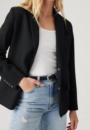 Kobieta w czarnym blezerze na białym prążkowanym topie na ramiączkach i jasnoniebieskich wysokich dżinsach z czarnym paskiem, ręka w kieszeni, neutralne tło.