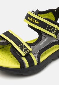 Svarta och gula sportsandaler med justerbara remmar och texturerade sulor. Har ett andningsbart material och en märkning som säger "VATTENVÄNLIG".