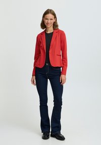 Röd figursydd kavaj med en enda knapp, bärs över en svart topp, ihop med mörkblå utsvängda jeans och svarta skor. Slät tygstruktur.