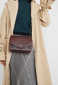 Burgundy crossover taske i tekstureret læder. Har en klaplukning og sølvfarvet hardware. Bæres over en mønstret nederdel og en beige frakke.