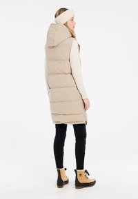 Gilet rembourré beige avec capuche, d'une longueur jusqu'aux genoux, associé à des leggings noirs et des bottes beiges, portant un bandeau en fourrure.