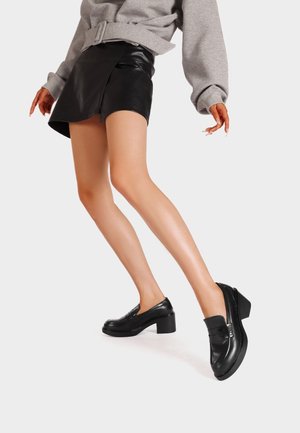 Kvinde iført sort lædermininederdel, grå sweatshirt med bælte og sorte hælede loafers, poserer med det ene ben strakt frem.