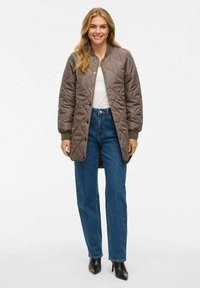 Glunderende vrouw met lang blond haar die een bruine gewatteerde lange jas, een witte top, blauwe jeans en zwarte hakken draagt, staande tegen een witte achtergrond.
