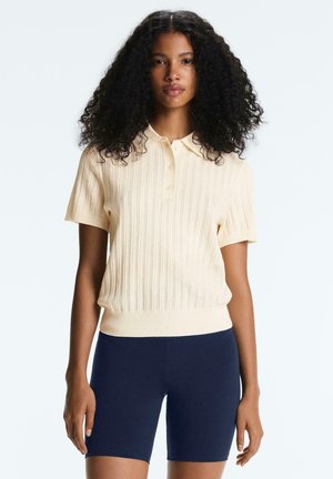 Donna con capelli ricci che indossa una polo in maglia color crema a maniche corte e pantaloncini aderenti blu navy, in piedi davanti a uno sfondo semplice.