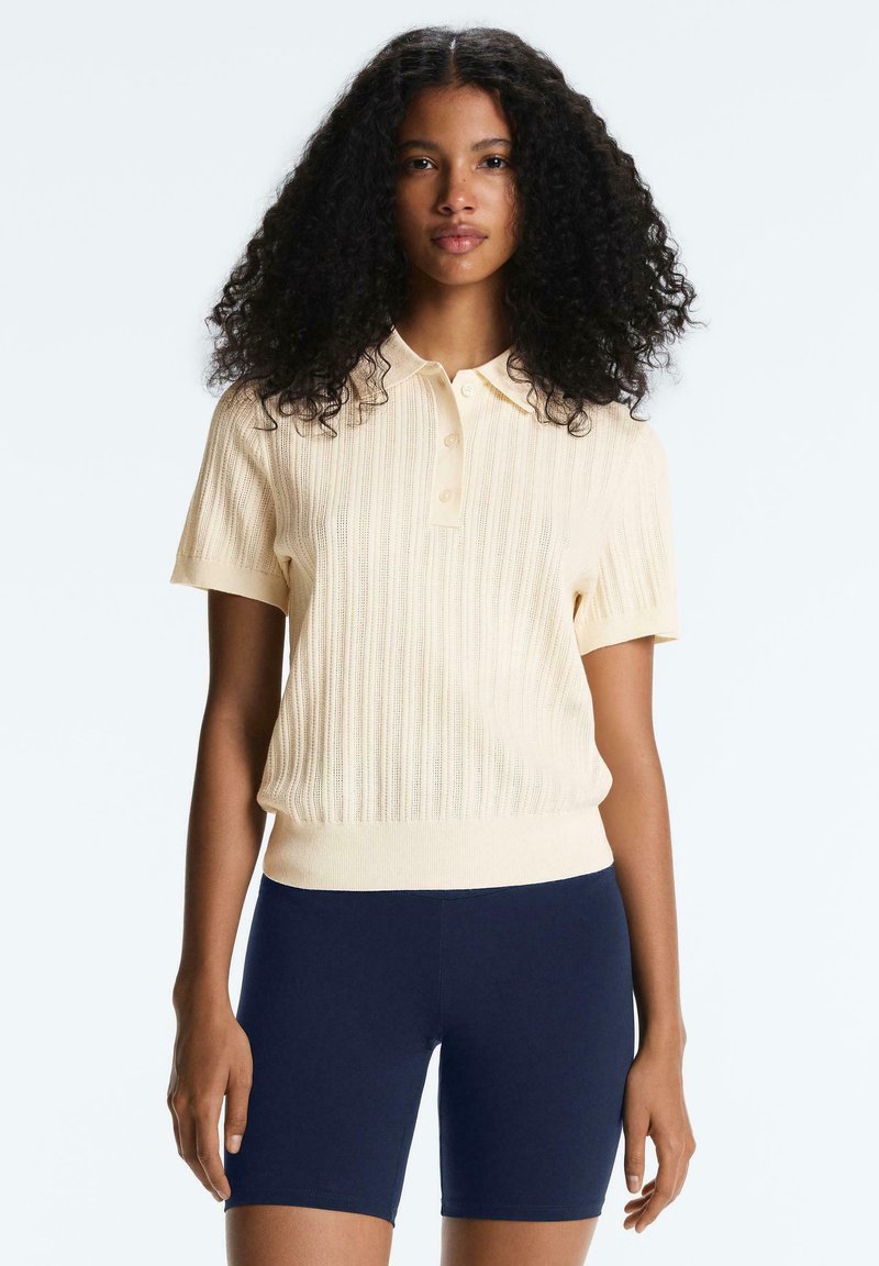 Woman with curly hair wearing a cream short-sleeve knit polo shirt and navy blue fitted shorts, standing against a plain background.