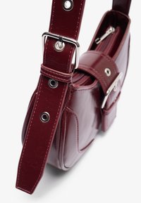 Bordeaux farbene Lederhandtasche mit verstellbarem Tragegurt, silberner Schnalle, Reißverschluss und einer Fronttasche, die mit einer silbernen Schnalle gesichert ist.