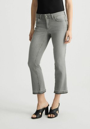 Jeans gris coupés avec une légère déchirure au bas, présentant un design classique à cinq poches et une fermeture éclair, associés à des sandales noires à talons.