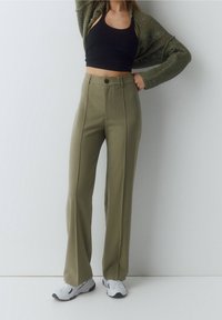 Olijfgroene wijde broek met een hoge taille, voorzien van een voorste knoopsluiting en verticale naden. Gepaarde met een zwarte crop top en witte sneakers.