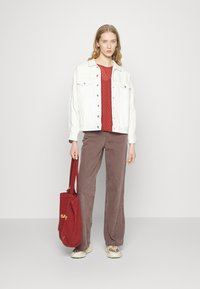 Jaqueta de ganga branca, blusa vermelha com bolinhas, calças largas de corduroy castanho e uma tote bag vermelha com "SAFE" em amarelo. Ténis bege.