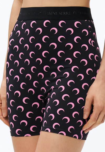 Czarne dopasowane szorty z różowym wzorem półksiężyców noszone na biodrach, ukazujące pas i górną część ud, z rękami spoczywającymi po bokach.