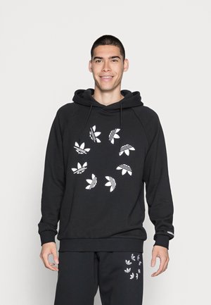 Felpa nera con una grafica bianca di foglie stilizzate e righe. Presenta un cappuccio con cordino e polsini a coste. Il modello indossa pantaloni abbinati.