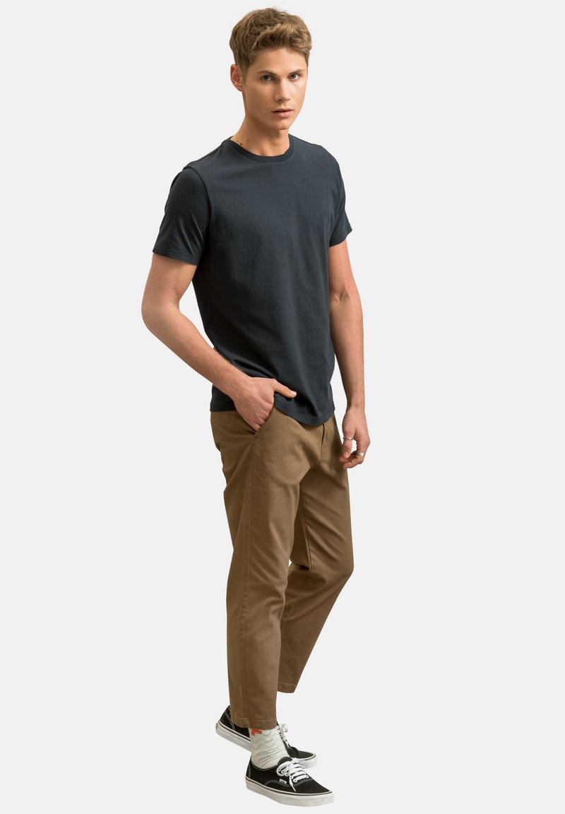 T-shirt blu scuro, pantaloni beige e sneakers nere con suole bianche; modello posizionato con una mano in tasca, sfondo neutro.