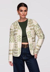 Femme portant un crop top vert, une veste beige et verte à motifs floraux avec des boutons, un jean foncé et une ceinture marron, posant avec la main sur la hanche.