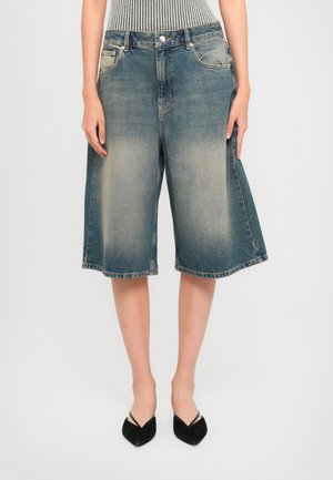 Slanke ben med falmede blå knælange denimshorts og sorte spidse hæle med tynde stropper på en lys baggrund.