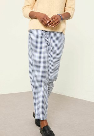 Person trägt einen beige gestrickten Pullover, blau-weiß karierten Hosen, schwarze Loafers, mit gefalteten Händen und einem blauen Perlenarmband.