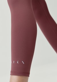 Bordové legíny vyrobené z elastického materiálu, s hladkým povrchom a malým bielym logom na leme. Dĺžka končí tesne pod kolenom.