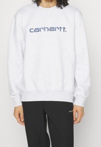 Persona che indossa una felpa grigia chiara di Carhartt con maniche lunghe e pantaloni neri a costine con del testo piccolo vicino alla tasca.