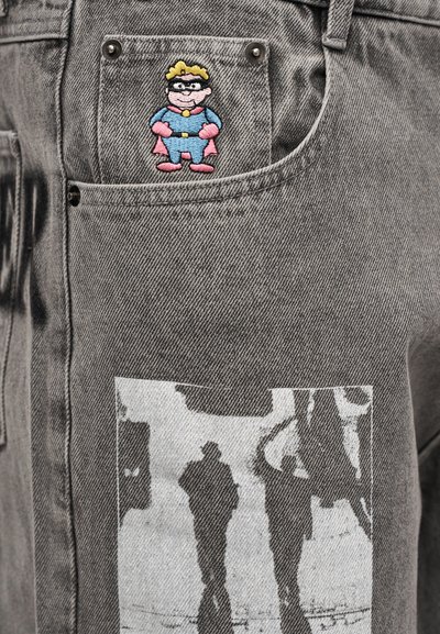 Γκρι τζιν από τζιν ύφασμα με ένα καρτούν-σουπερήρωα στο μπροστινό τσεπάκι και ένα ασπρόμαυρο φωτογραφικό τύπωμα στο μηρό.