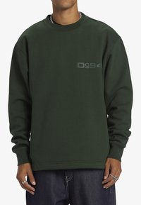 Grön sweatshirt i tjockt material, med ribbad rund halsringning, långa ärmar och en stiliserad grå logo på bröstet. Avslappnad design.