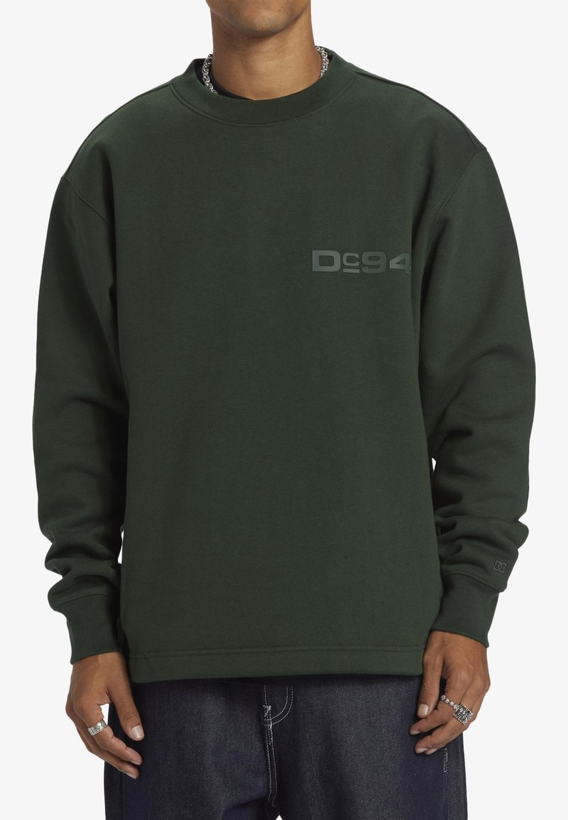 Grön sweatshirt i tjockt material, med ribbad rund halsringning, långa ärmar och en stiliserad grå logo på bröstet. Avslappnad design.