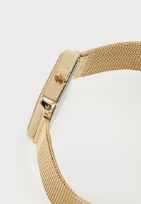 Montre en métal doré avec un boîtier rectangulaire, un bracelet en maille, des accents en cristal clair et une couronne latérale pour les réglages.
