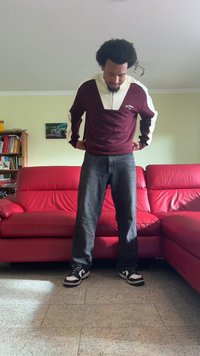 Bordó és bézs zipzáras pulóver fehér részletekkel, sötét farmernadrággal és fekete-fehér sportcipővel párosítva, egy nappaliban állva.