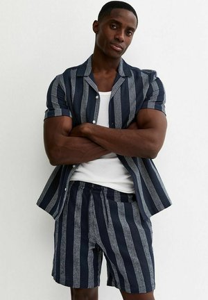 Uomo in piedi con le braccia incrociate che indossa una camicia a righe verticali blu navy e bianche a maniche corte e pantaloncini abbinati sopra una canottiera bianca.