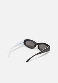 Svarte og hvite solbriller med vinklede glass. Svart front og armer, hvit detaljering på venstre arm. Strukturert overflate og mørke glass.