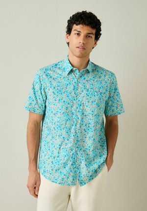 Mand iført en turkis kortærmet skjorte med knapper og et lille blomsterprint samt off-white bukser, stående mod en ensfarvet baggrund.