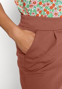 Bruna, strukturerade byxor med elastisk midja och sidofickor. Tyget är slätt med subtila veck som ger dimension.