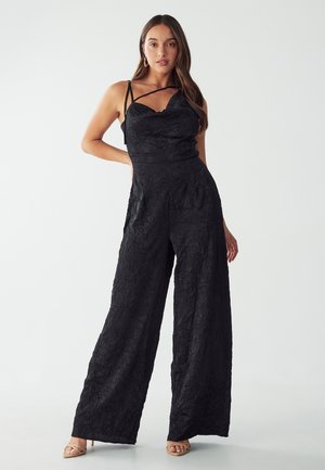 Black jumpsuit with a textured finish, featuring spaghetti straps and a scalloped neckline. Wide-legged design with relaxed fit.
