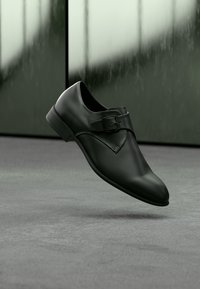 Μαύρο δερμάτινο παπούτσι με λεία υφή, στρογγυλεμένη μύτη, διακοσμητική αγκράφα στο πλάι και χαμηλό τακούνι, τοποθετημένο πάνω σε γκρι επιφάνεια.