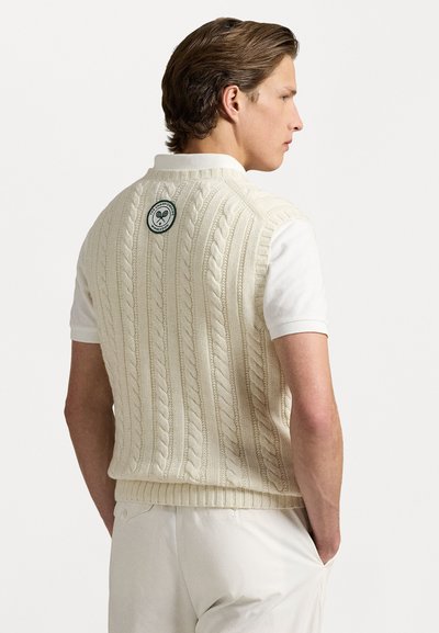 Chaleco de punto color crema con un patrón de trenzas, dobladillo acanalado y una insignia redonda en la parte posterior. Lleva puesto sobre una camiseta polo blanca.