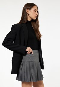 Sort blazer over en tætsiddende sort højhalset trøje, parret med en grå plisseret nederdel med pincetstriber og sidelommer.