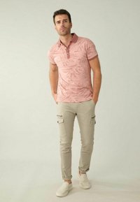 Hellrosa Polo-Shirt mit einem Blättermuster und einem kontrastierenden Kragen. Kombiniert mit beigen Cargo-Hosen und weißen Slippern.