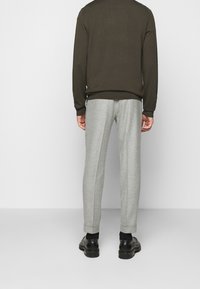 Uomo che indossa un maglione marrone a maniche lunghe, pantaloni grigi chiari su misura con risvolto, calzini neri e scarpe di pelle nere, in piedi contro uno sfondo bianco.