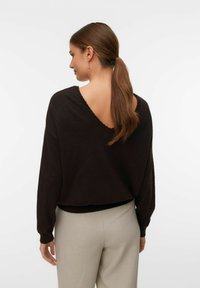 Maglione marrone, dal taglio ampio con un profondo scollo a V sul retro, maniche lunghe e orlo a costine, abbinato a pantaloni chiari.