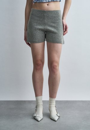 Kvinde iført grå strikkede shorts med små hvide prikker, hvide midt-lange sokker og hvide spidse sko med spænder, stående på grå gulv.