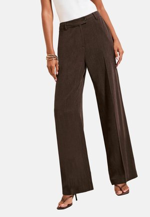 Femme portant un pantalon marron foncé taille haute à jambes larges, associé à un top blanc sans manches et des sandales marron à talons.