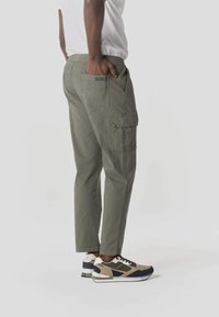 Hombre de pie con la mano en el bolsillo trasero de unos pantalones cargo color verde oliva y luciendo zapatillas beige, verdes y negras sobre fondo blanco.
